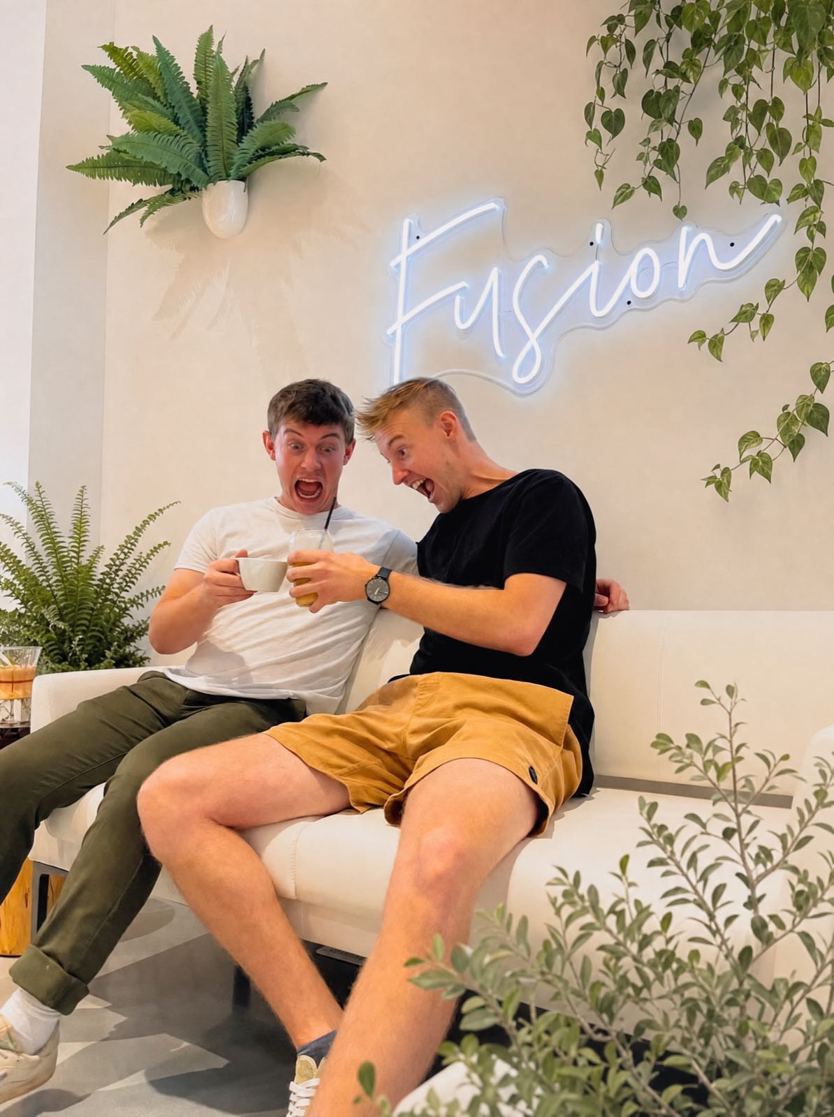 Two friends laughing on the cream sofa beneath the blue neon Fusion sign, clinking a coffee cup against an iced drink, surrounded by ferns and trailing greenery.